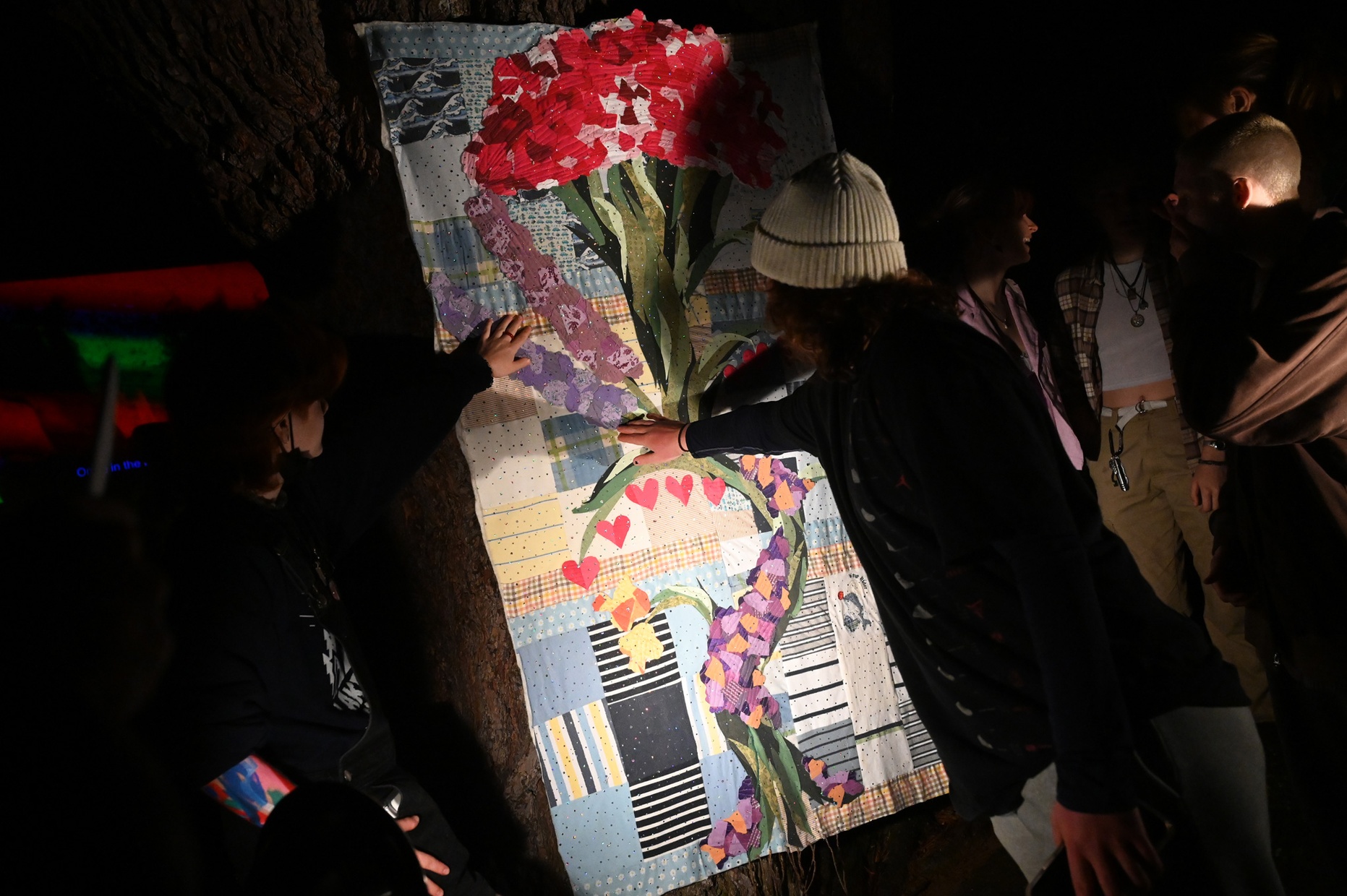 In the dark, people gather around to touch a quilt lit by spotlights.