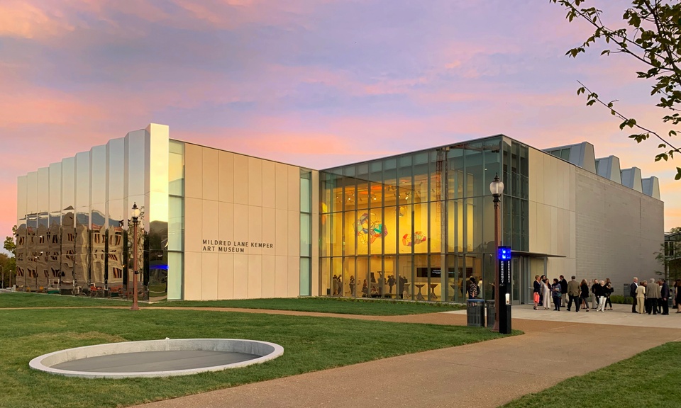 Kemper Art Museum backed by a pink and purple sunset. The interior is lit and a Tomas Saraceno sculpture can be seen suspended from the ceiling of the lobby.