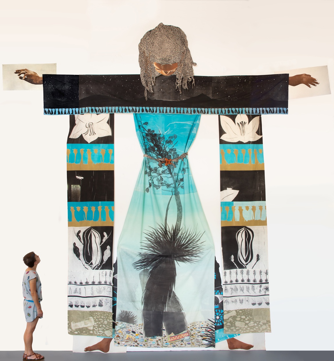 An adult stands in the left foreground, looking up at a large, flatly-collaged humanoid being who stands with their head down and arms and legs outstretched. 