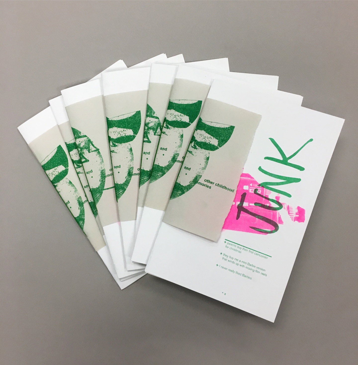 A group of several copies of the same zine. Each zine's spine is wrapped with a green-on-gray print depicting imagery and the words "and other childhood memories." The cover of the zine is white with pink imagery and green lettering. The word "JUNK" is big and handwritten. The following list appears below it in smaller text: "+ parents buy their first camcorder for christmas" "+ they buy me a mini Barbie version that winds up with moving film reels" "+ I never really liked Barbies"