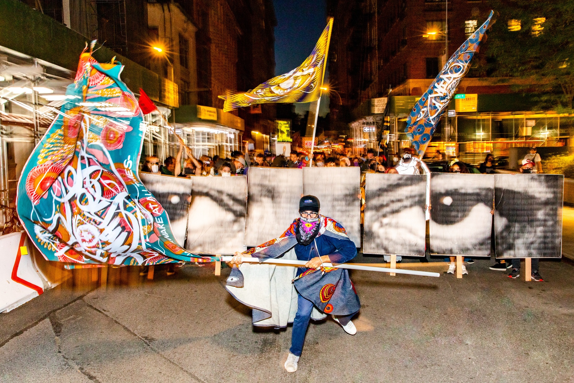 A photograph of a person in the street at night, waving a colorful flag. Behind them is an out of focus crowd and a banner with a photograph of eyes.