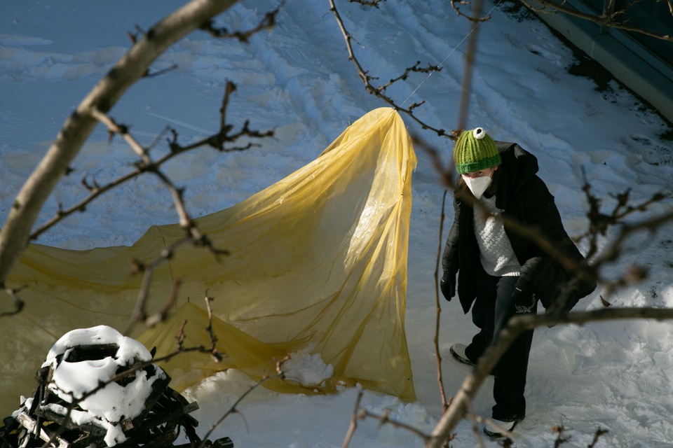 Outdoor photograph of a person standing in the snow to the right of a large yellow piece of fabric draped from a tree.