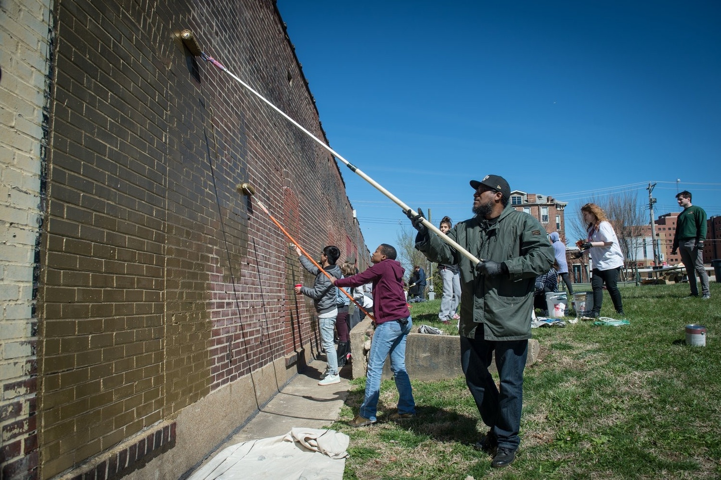 Two people paint a wall gold with extended rollers, while other people paint in the background. 