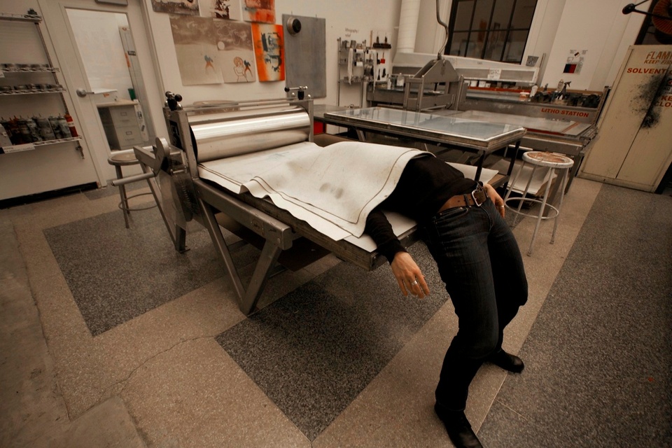 Photo of a person lying under white fabric on a press, the top of their torso covered, with legs sticking out and feet on the ground.