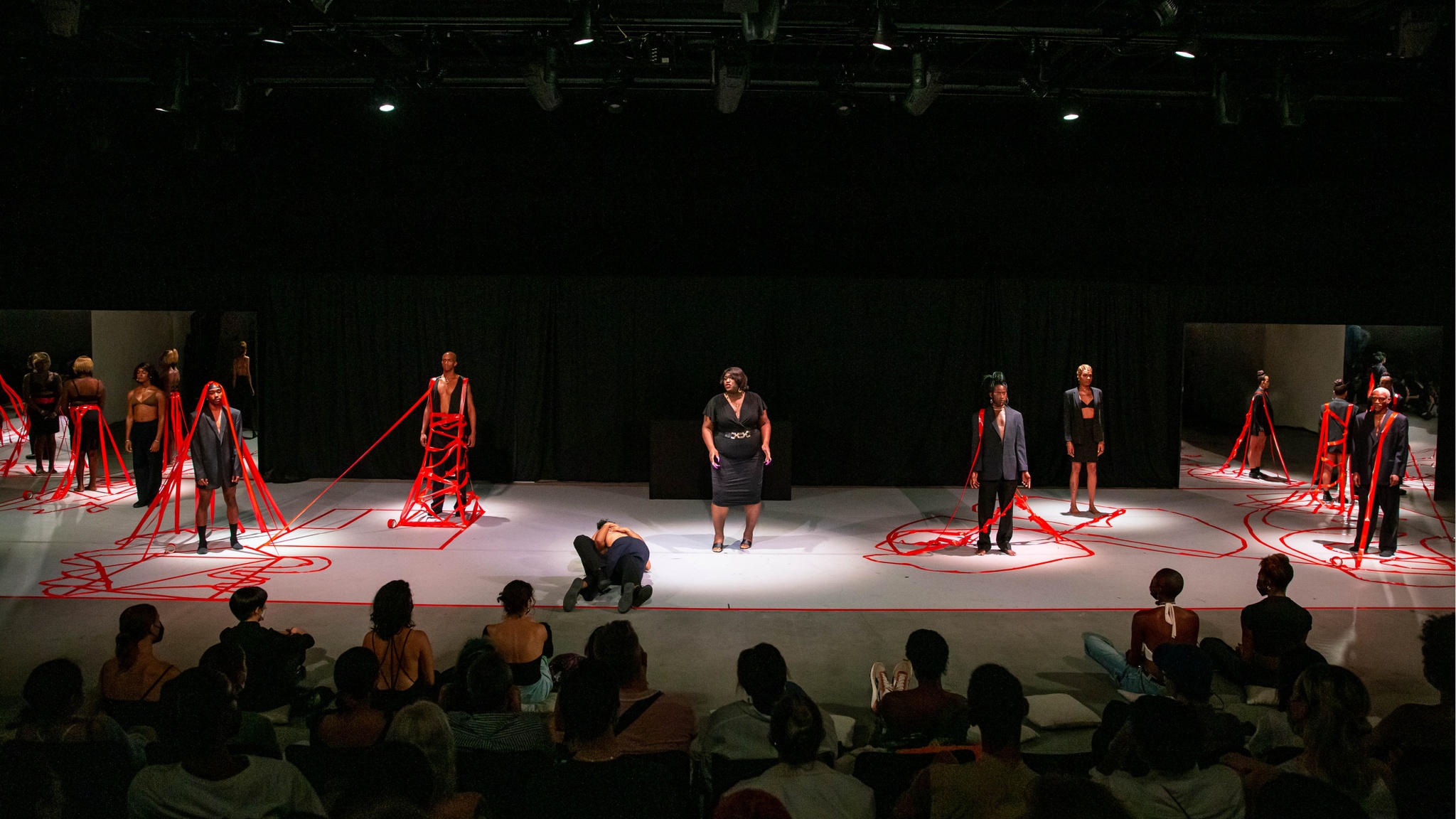 Nine Black trans performers stand on a stage wearing black suits and dresses in front of an audience. On the floor of the stage, two performers embrace as they roll together across the floor. Six of the standing performers have red tape reaching up from the ground to hold them in place.