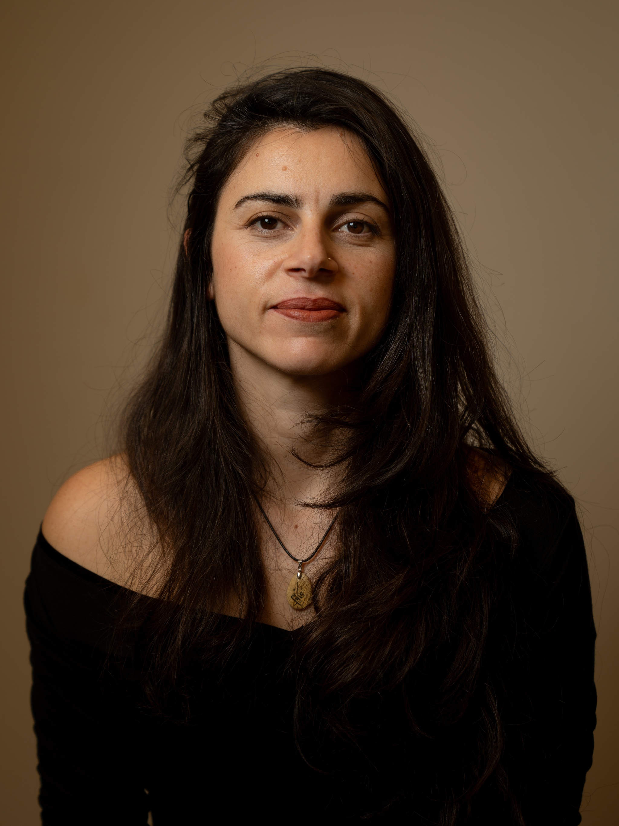 A portrait of Samaa Wakim against a neutral dark brown background. Samaa has long dark brown hair that falls below her shoulders. She wears a shoulderless top and looks directly at us, smiling slightly. 