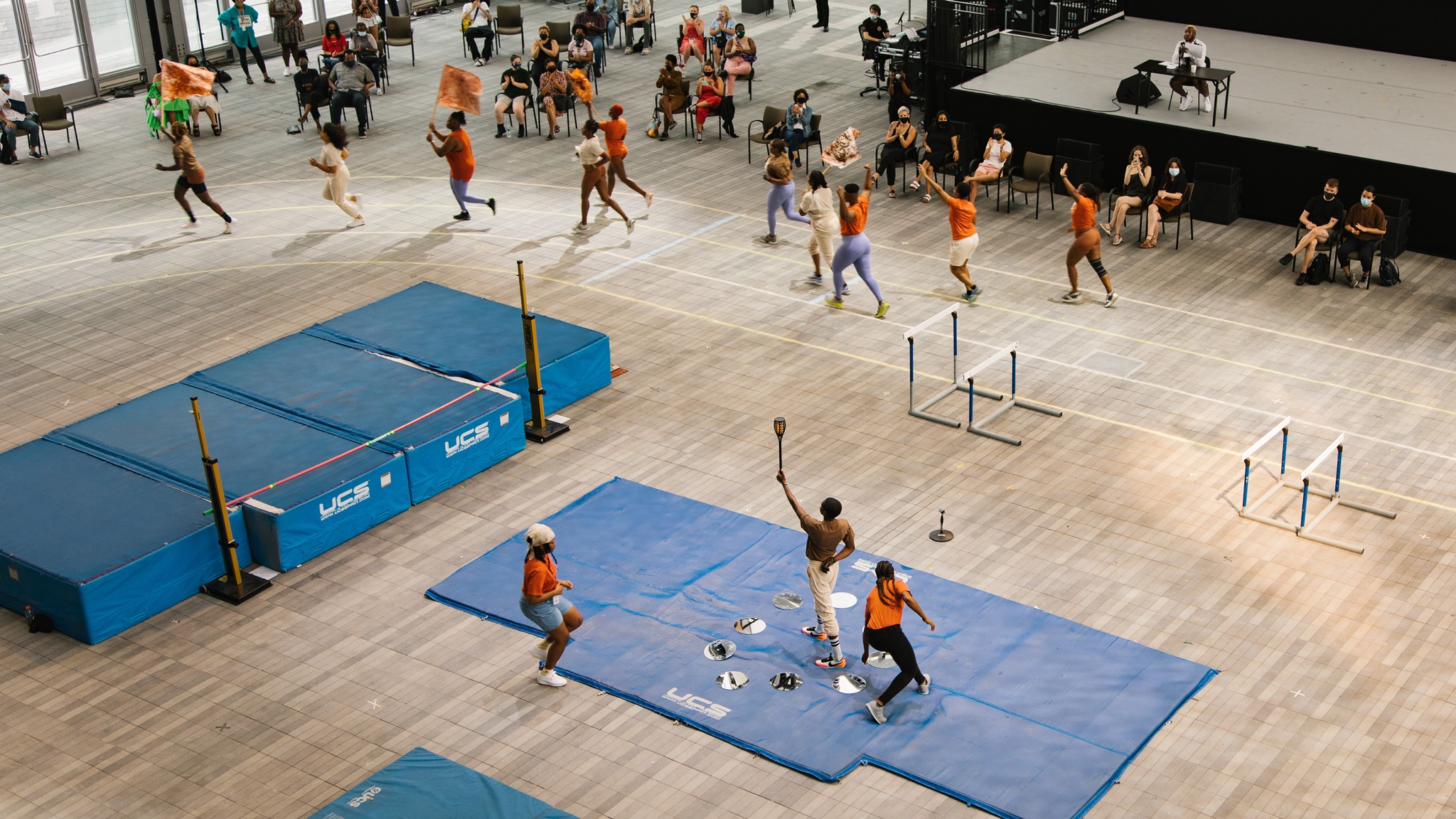 Blue athletic mats laid out in an open performance space. Athletes are running around a track that encircles the mats. 