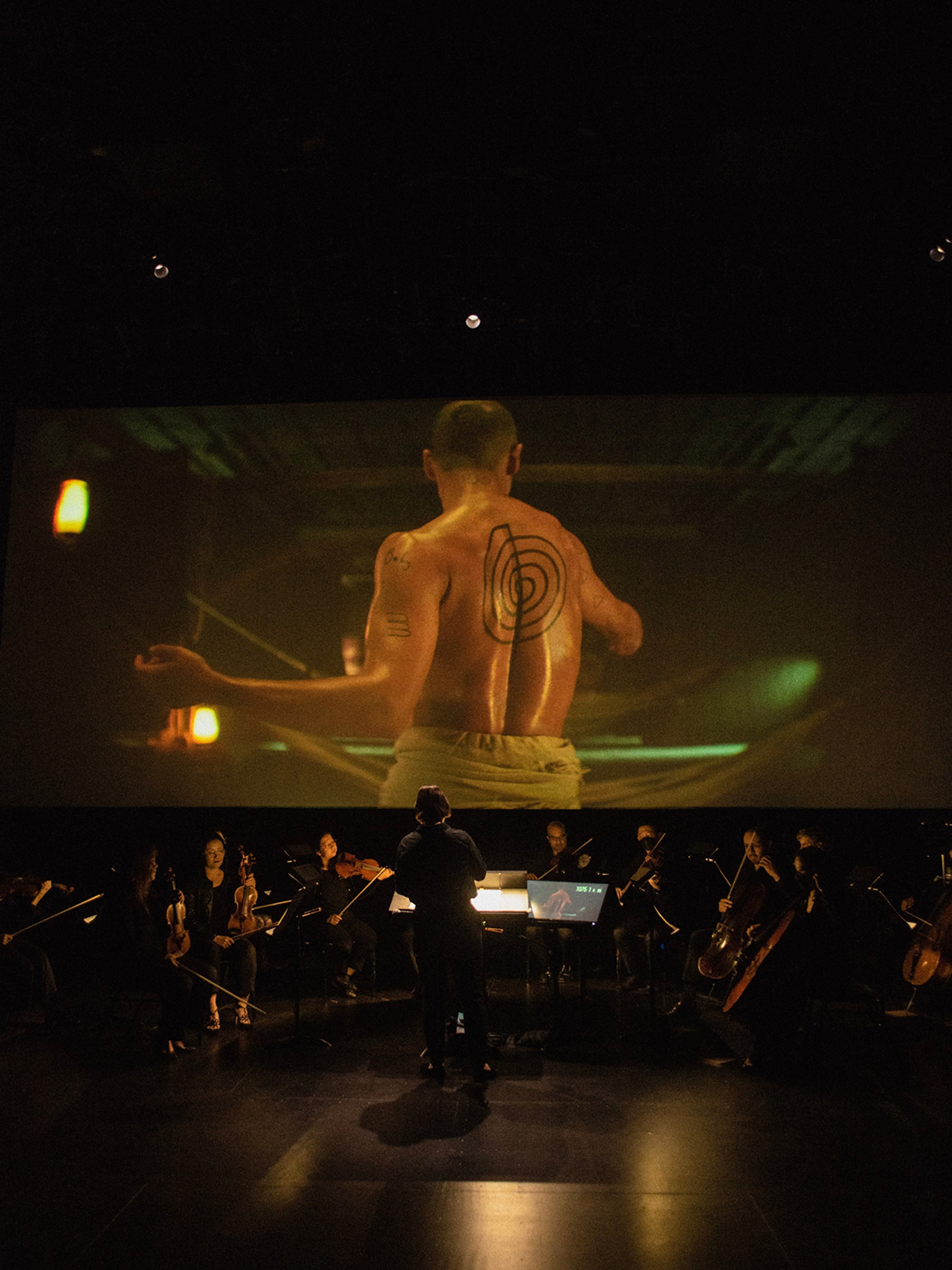 A small orchestra plays in a dark theater space in front of a film screen. On the screen is projected a scene from Wu Tsang's Moby Dick: a shirtless person with a tattooed muscular back stands with arms out to the side.