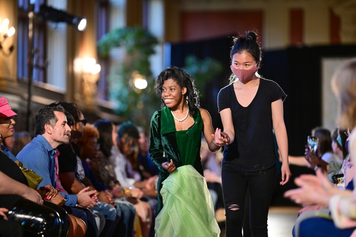 A model in a green velvet dress walks down a runway hand in hand with a student designer