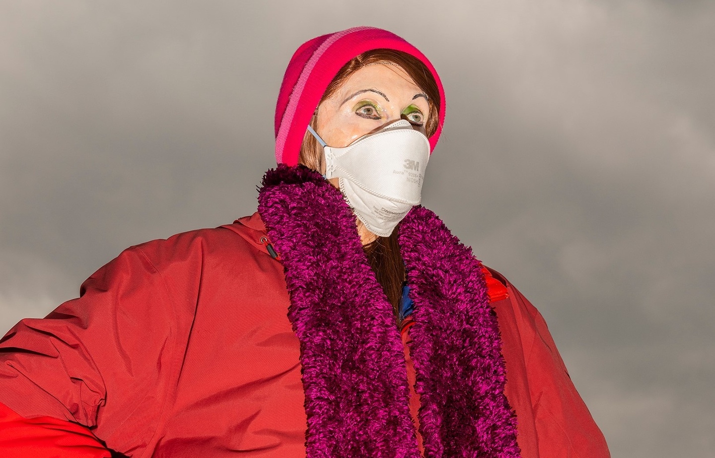 A brightly lit shot of Linda Leven wearing a white mask, red beanie, red jacket and a magenta boa with a backdrop of a downcast sky
