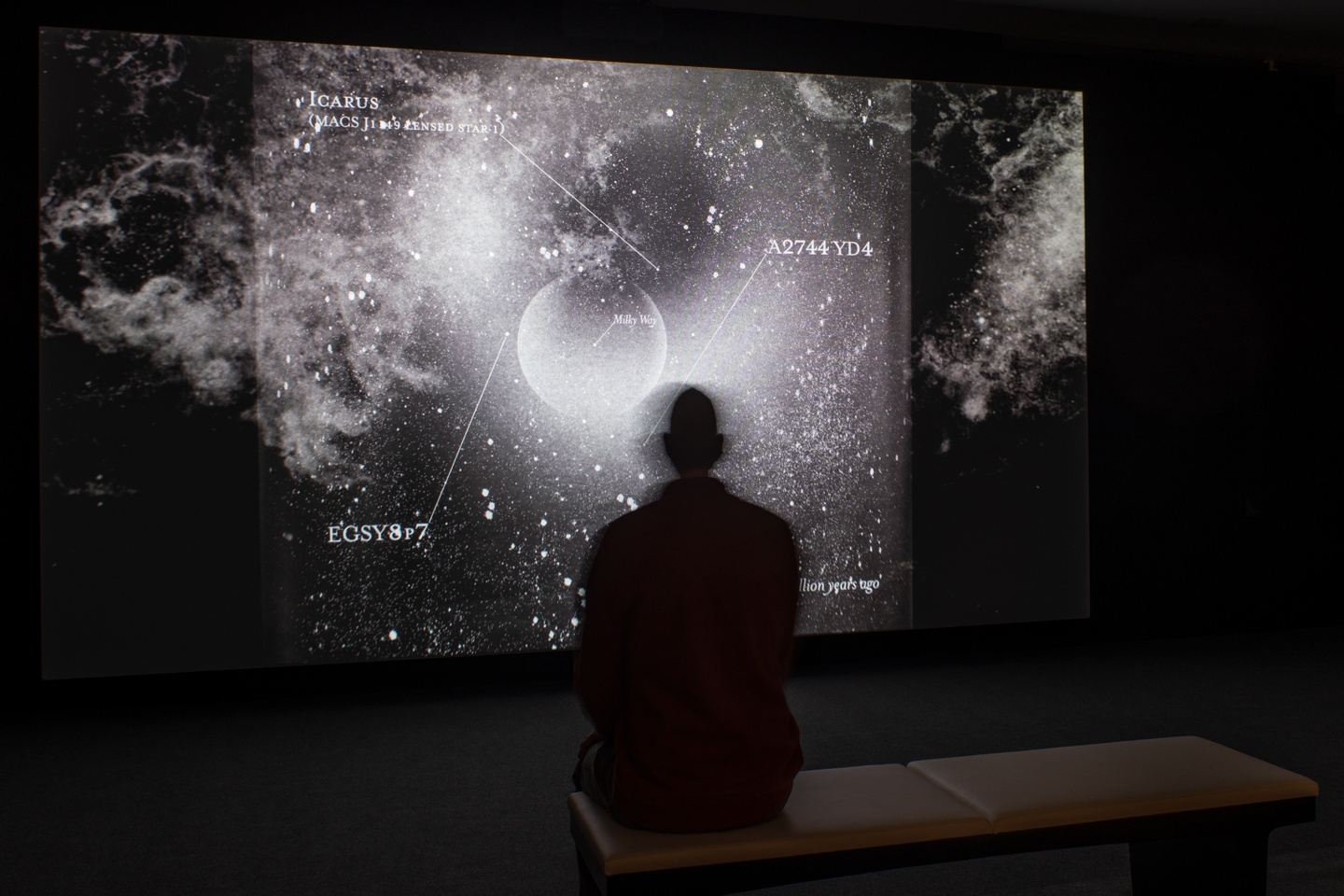 Mostly black-and-white video still of a person seated on a dark tan bench, their back to the camera, staring at a screen displaying a view of the galaxy, with the Milky Way, Icarus, and a couple of other elements pointed out.