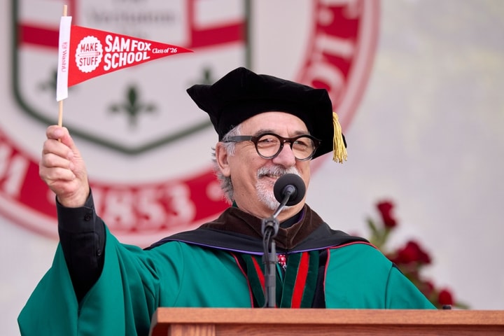 A man wearing regalia stands at the lectern while waving a Sam Fox School pennant.
