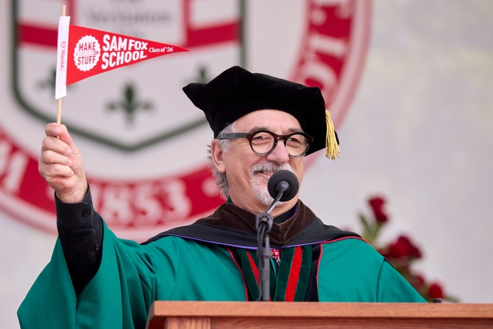 A man wearing regalia stands at the lectern while waving a Sam Fox School pennant.