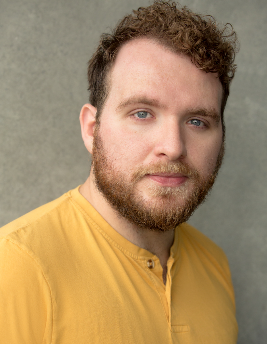 A headshot of actor Hughie O'Donnell, who turns his head to look directly at us. Hughie is white and has light brown curly hair and a beard. He wears a yellow tshirt. 