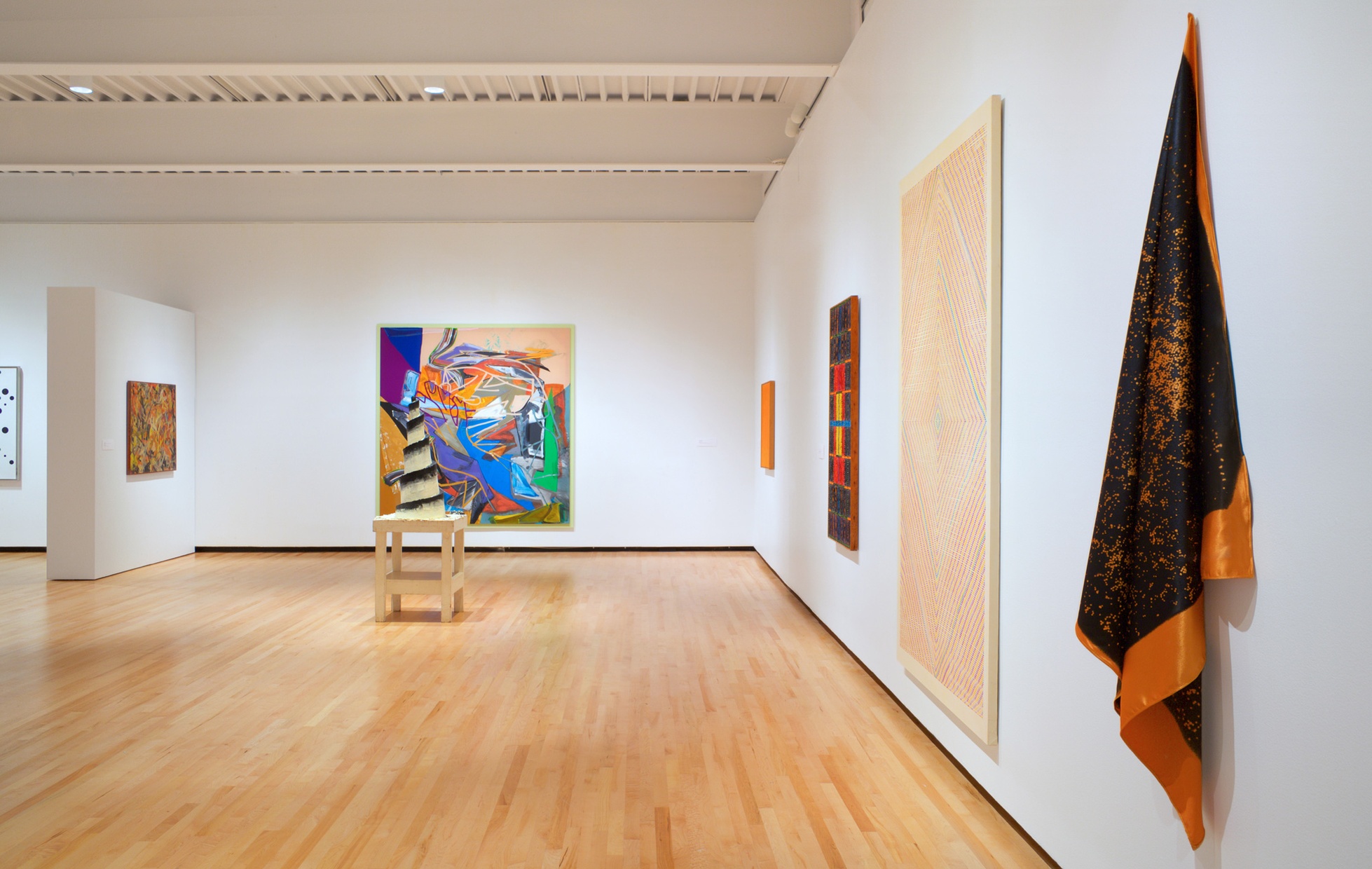Artworks with bright, warm colors hang on white walls of a large room with a sculpture in the center of the warm wood floors.