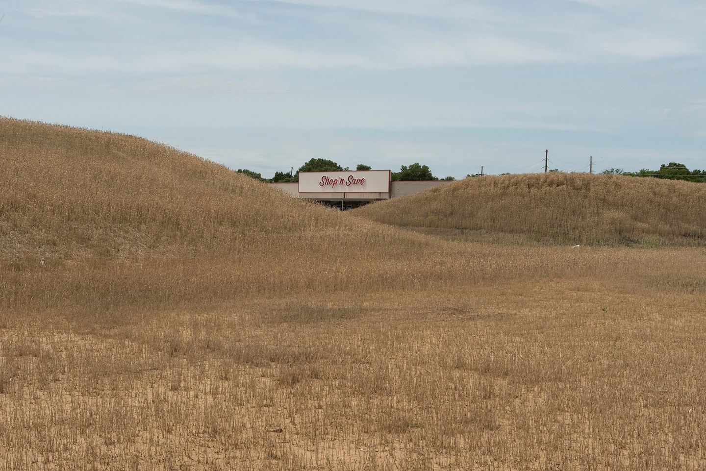 Beyond two golden mounds/hilltops, the sign "Shop 'n Save" in a red calligraphic brush font, on the façade of an obscured storefront, centered in the photograph. The sky is a light blue, the clouds washed away.