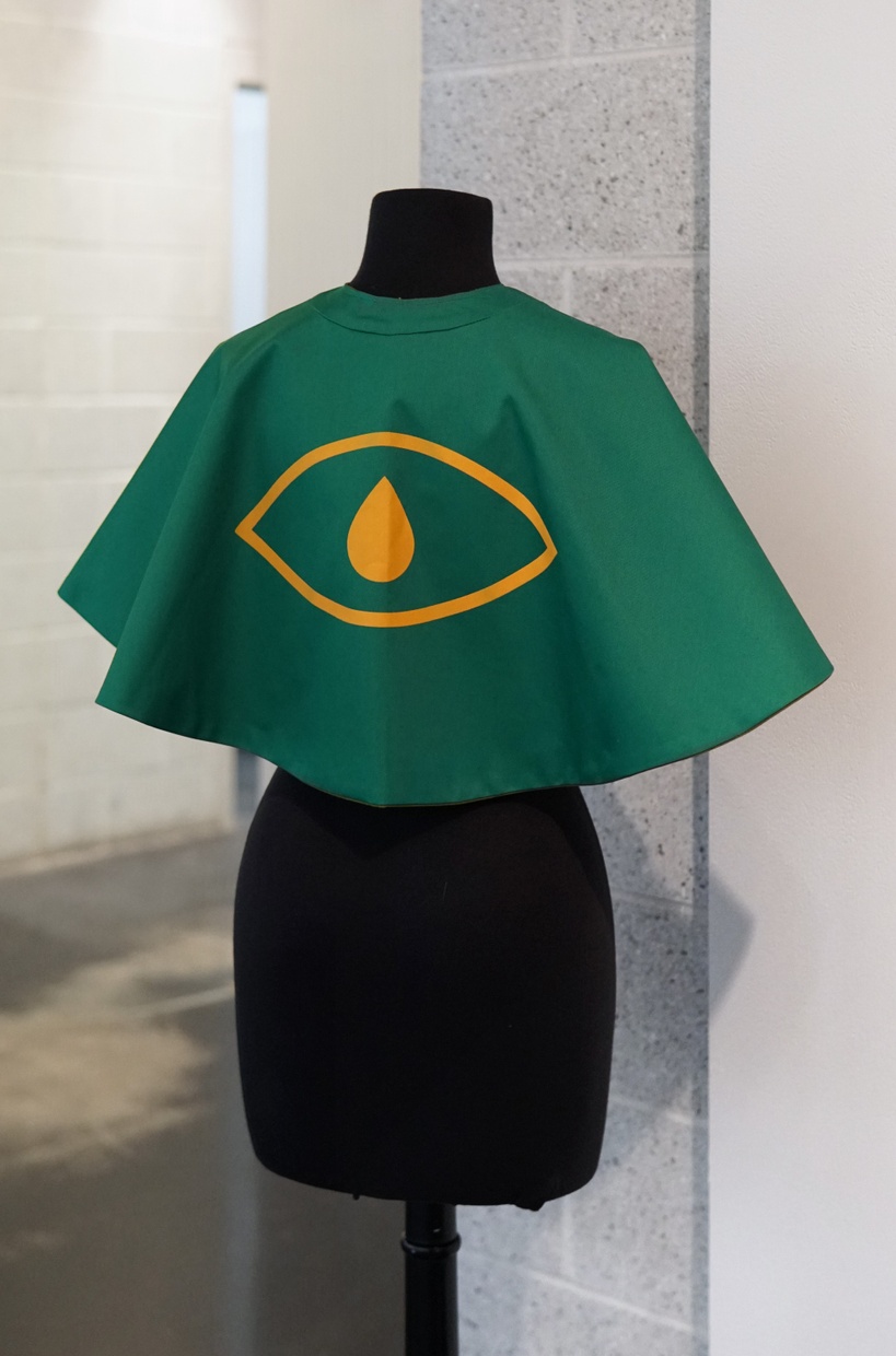 A short, green cape with a simple yellow outline of an eye on the back, and a solid yellow teardrop-shaped iris within the eye outline.