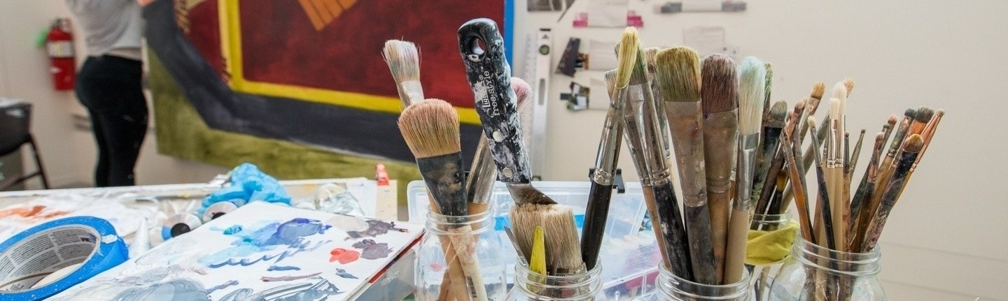 Artful shot of jars of brushes on a table next to a student working on a large-scale painting.