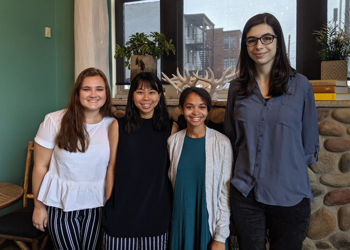 Four people stand in a row in front of a window, smiling at the camera.