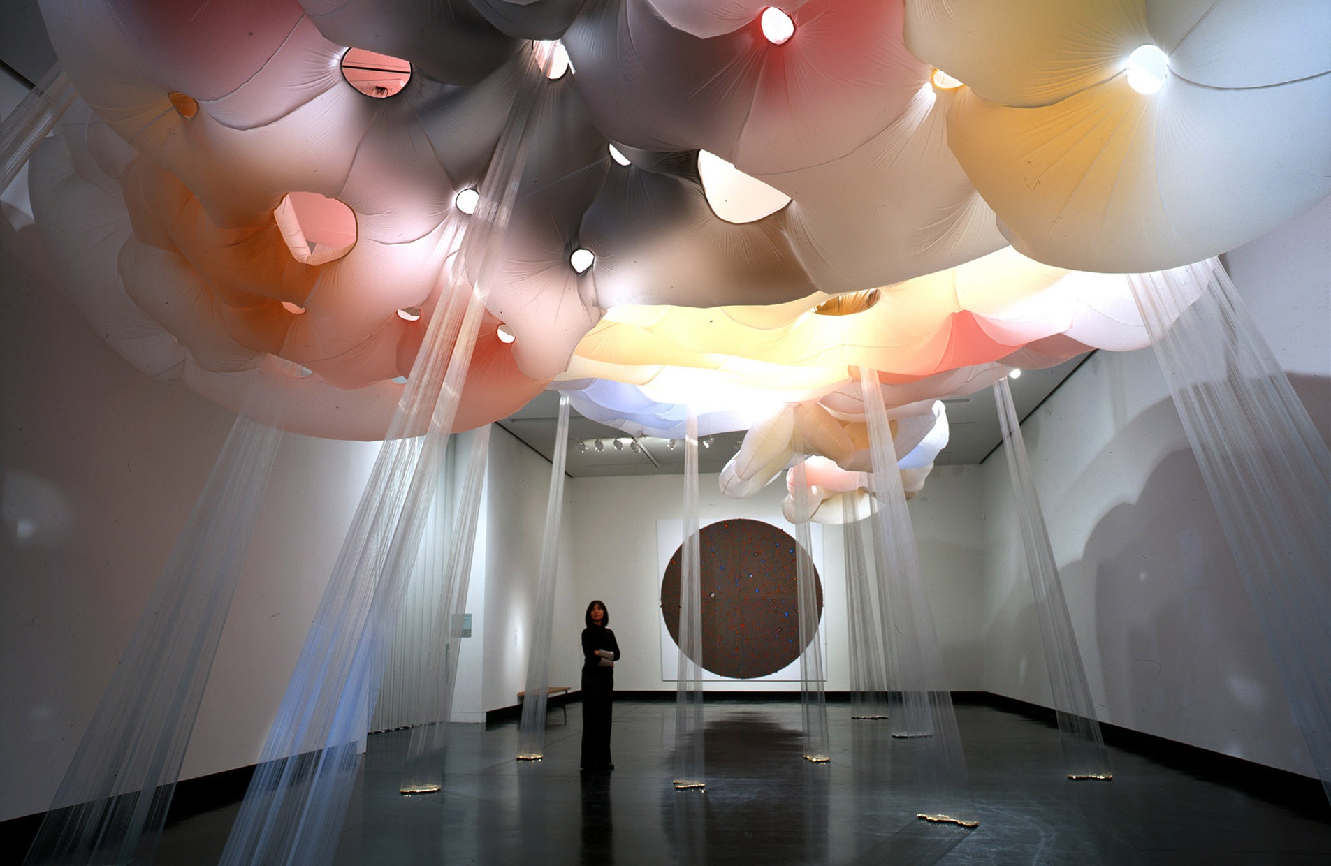 A woman stands in a gallery looking up at a red, yellow, black, and white cloud-like sculpture on the ceiling with rays extending to the floor.