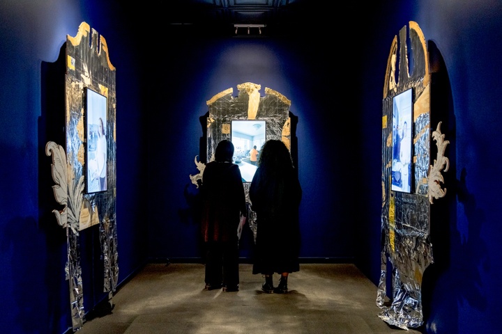 Two silhouetted figures stand in a small, dark blue room with three tvs installed inside of wooden frames, which are shaped like arched door lintels and covered in metallic film with gold trim.