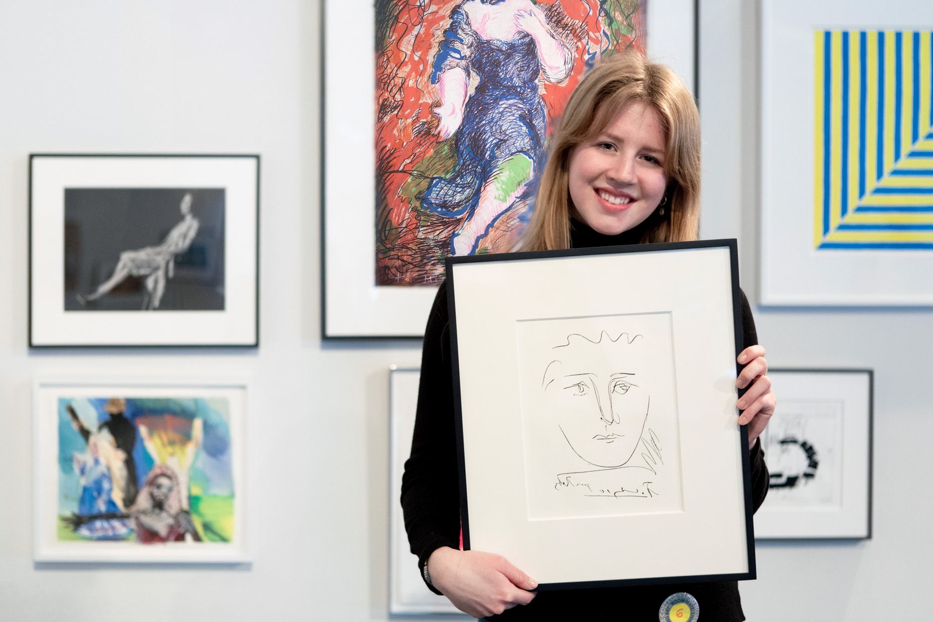 A woman is smiling holding a framed print by Picasso, more framed artwork hangs on the wall behind her.