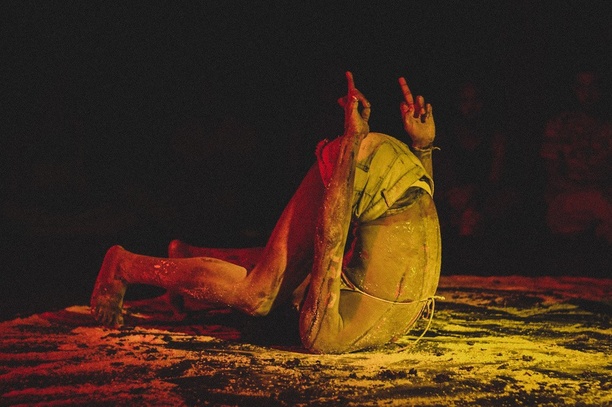 A person wearing a bikini top and short jeans is contorted, touching the floor only with the back of the head and the tip of the feet. The person's butt is pointing upward, as are the two arms that vertically lift middle fingers. The marks on the white powder over the dark floor seem to keep traces of intense movement. The person is bathed in red and yellow lights