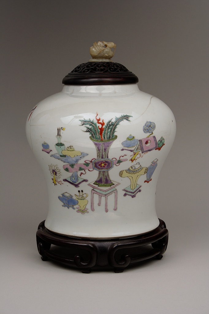 A white jar, with black lid and base, depicts colorful paintings of different shaped vases with the center one being the largest and full of peacock feathers.