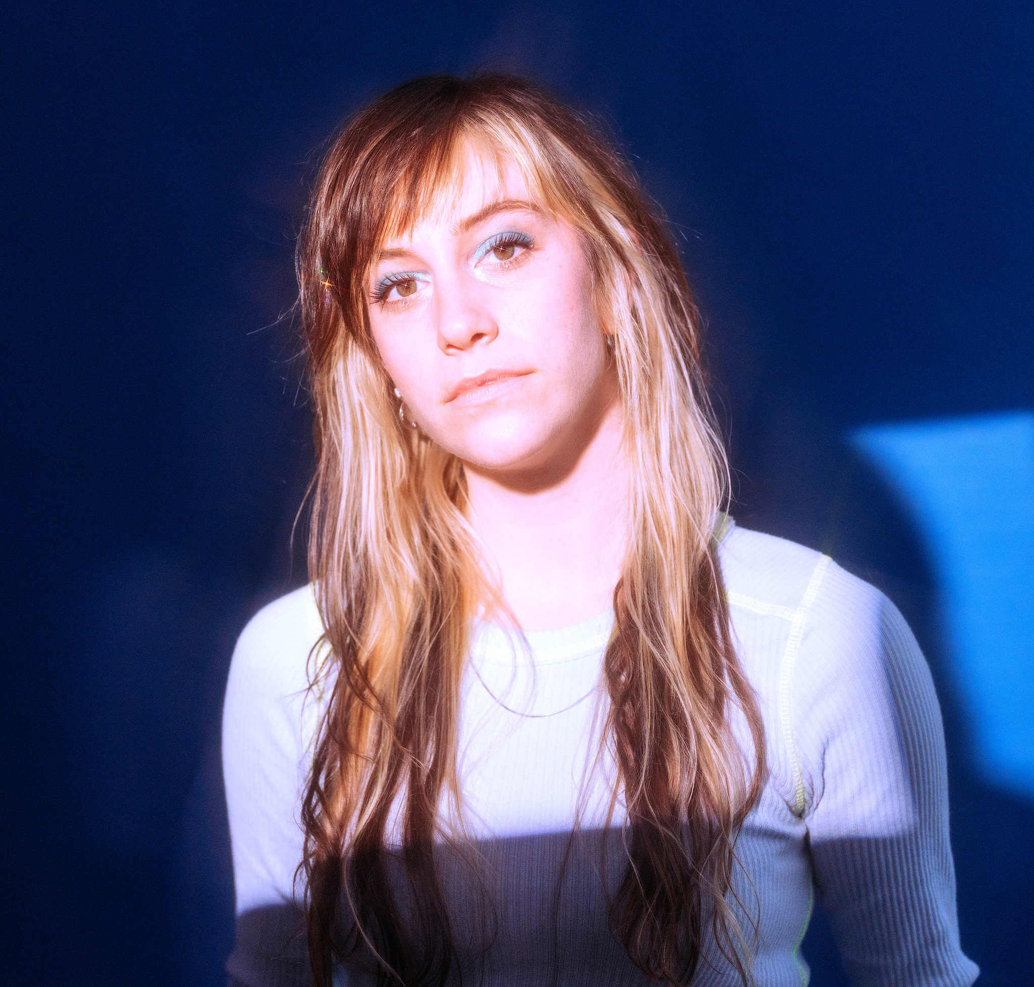 A portrait of Ellen Winter against a blue background. Bright light falls across Ellen's face as if she is facing an open window. She has long light brown hair that falls down beneath her shoulders. She looks directly at us. 