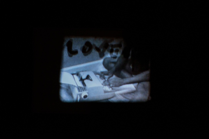 Grainy black-and-white film projector still of a person sitting cross-legged on the floor, typing on a typewriter. Handwritten text mars the film, in this frame, the word "LOVE."