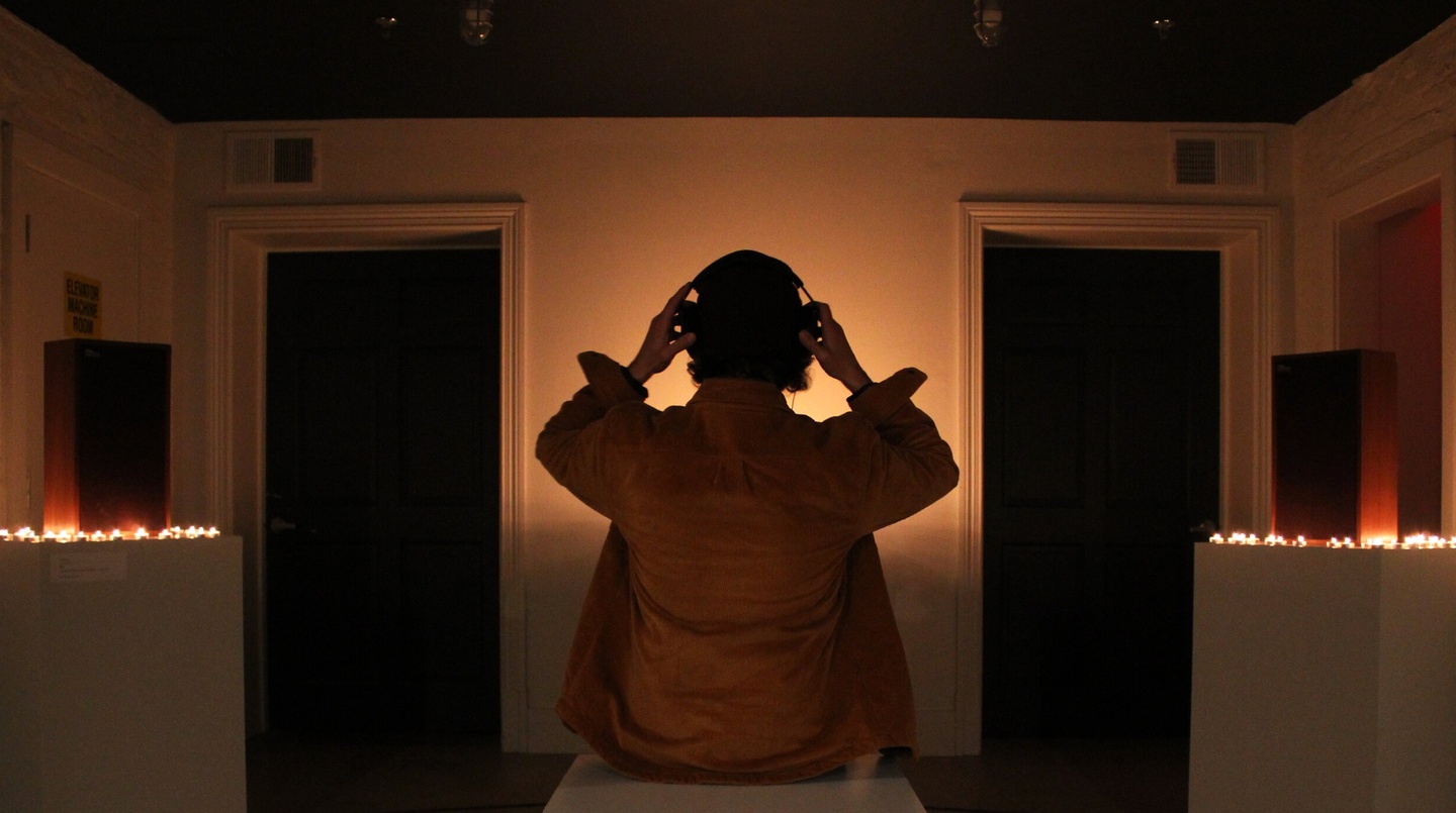 Person in a dimly lit room, standing with their back to the camera, hands on their head and wearing headphones. Candles are lit on pedestals to the left and right.