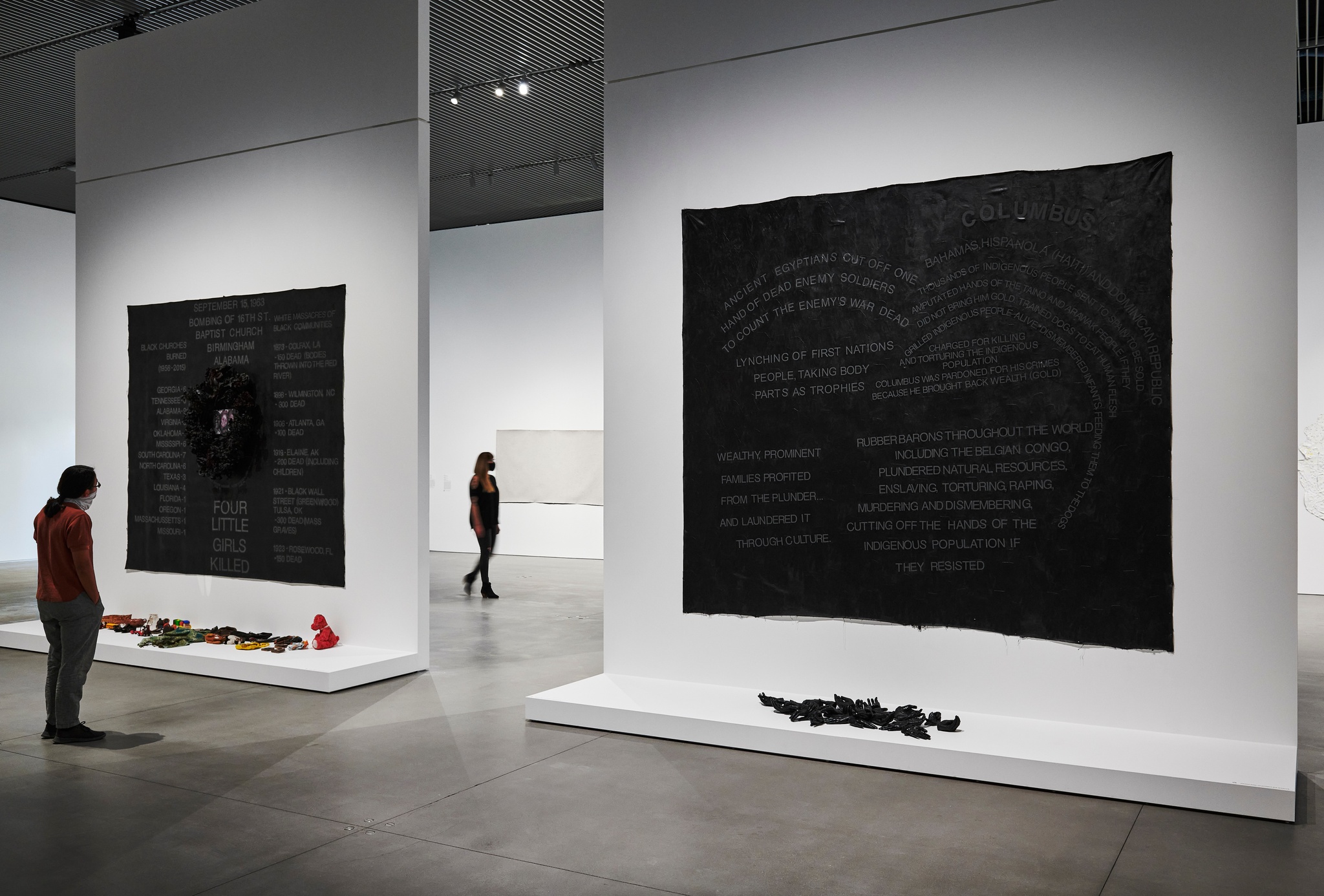 An installation view of two thematic paintings in the exhibition Howardena Pindell: Rope/Fire/Water. A gallery visitor looks at two square black paintings on adjacent walls while another visitor crosses the space in the background. The black paintings have text applied to their surfaces and objects arranged at their bases.