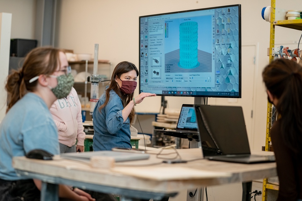 Instructor demonstrates 3D modeling software on a large screen to a group of people with laptops.