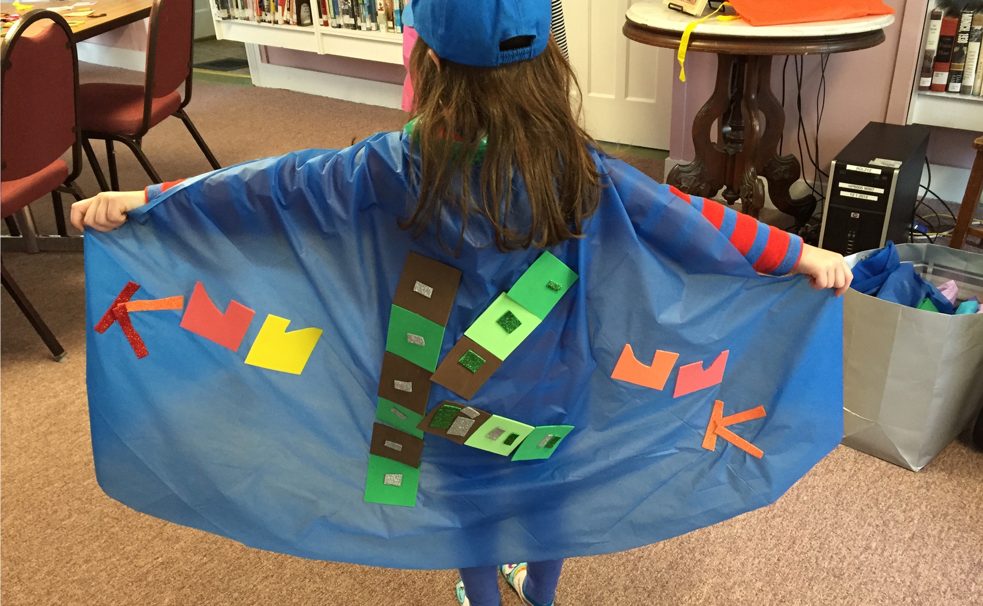 A young child, facing away from the camera, displays a blue cape with the letter K made from squares of colorful paper.