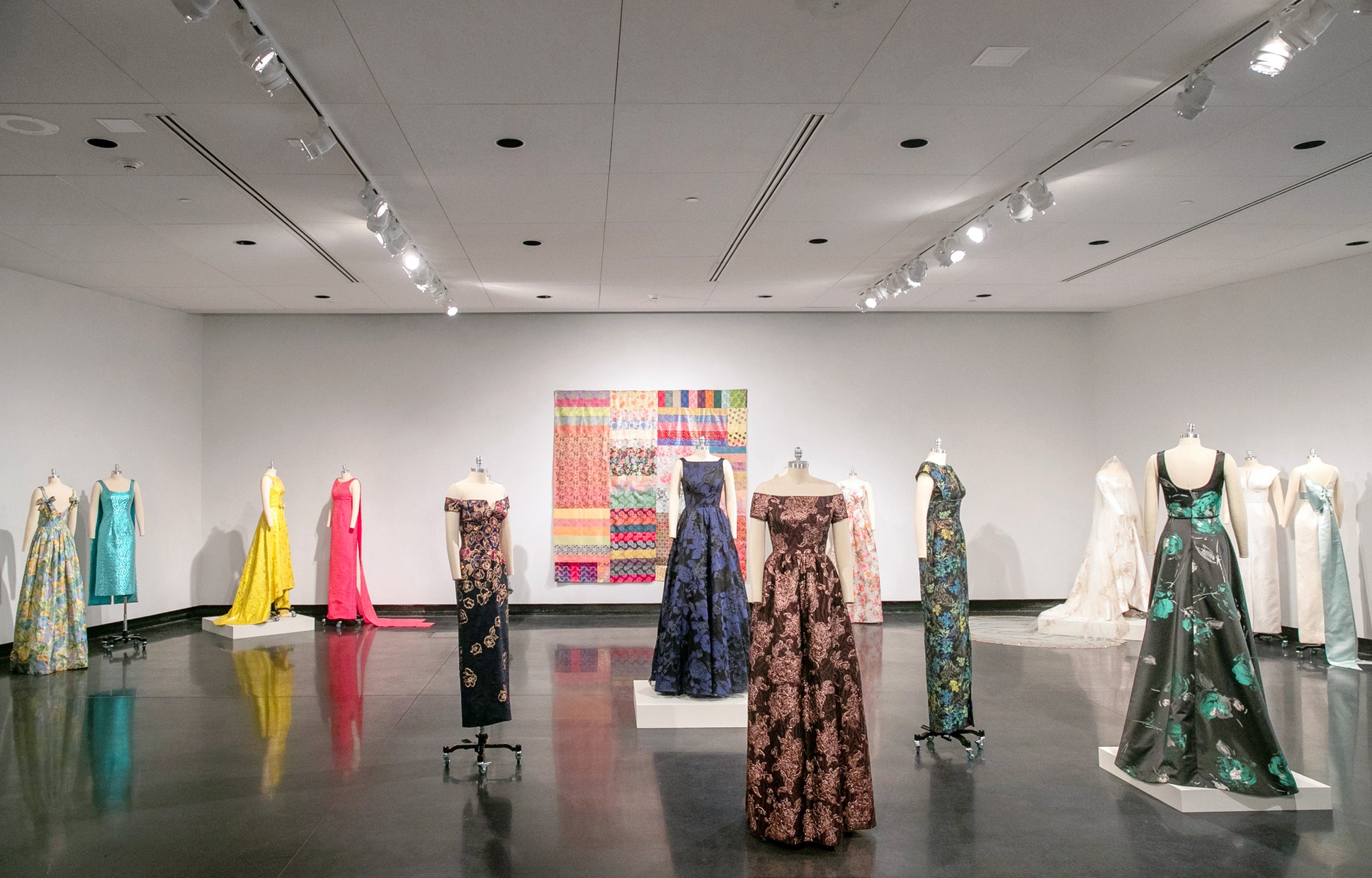 Multiple mannequins wearing colorful gowns placed throughout a room with a quilt hanging on the back wall.