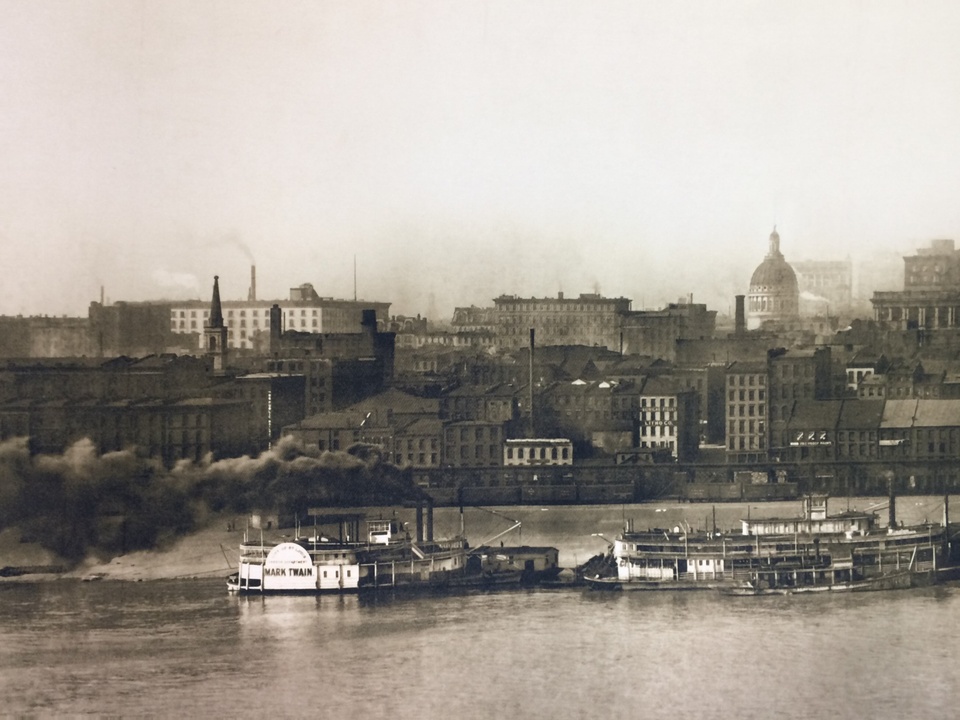 Archival, sepia-toned photograph of the old St. Louis waterfront, with barges on the water and buildings in the background.