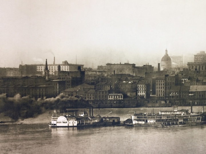 Archival, sepia-toned photograph of the old St. Louis waterfront, with barges on the water and buildings in the background.