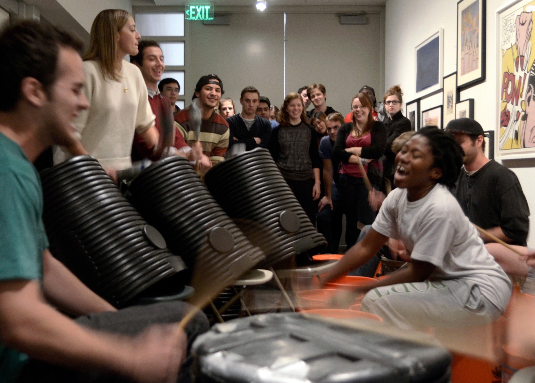 A group of young people bang on different types of cans like drums while a crowd watches from behind them.