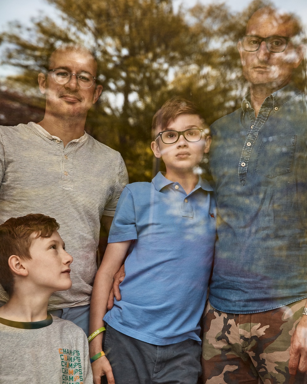 A photograph of a light-skinned family, two adult males wearing glasses and two boys, looking out a window with the reflection of a tree on it.
