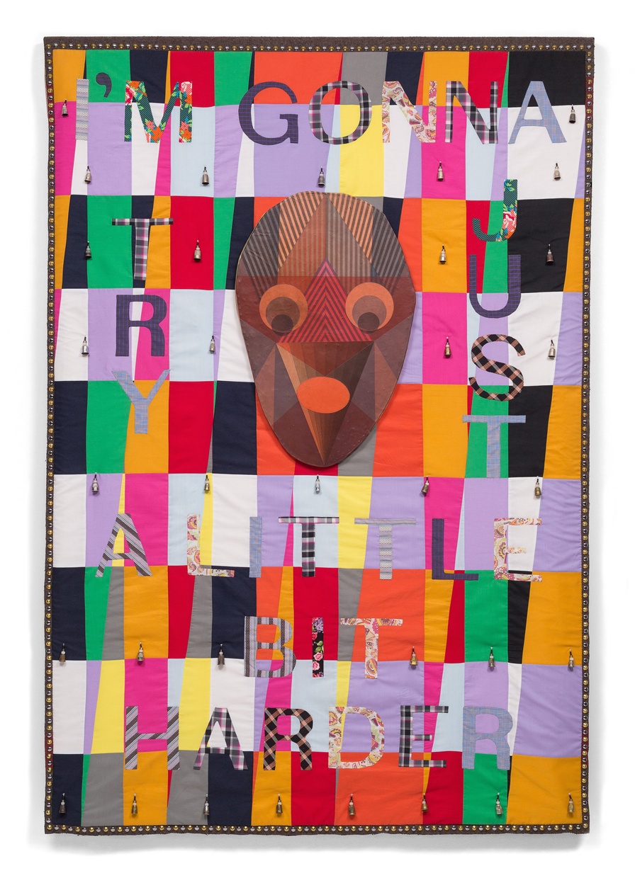 A colorful patterned quilt with a brown mask hanging in the upper middle and quilted text “I’M GONNA TRY JUST A LITTLE BIT HARDER” throughout the quilt.