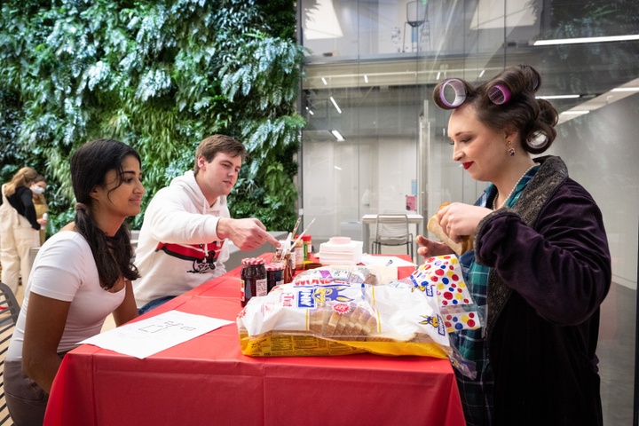 Two people sit at a table covered in a red tablecloth. A person in oversized hair rollers and a dressing gown stands behind the table making PB&J sandwiches.