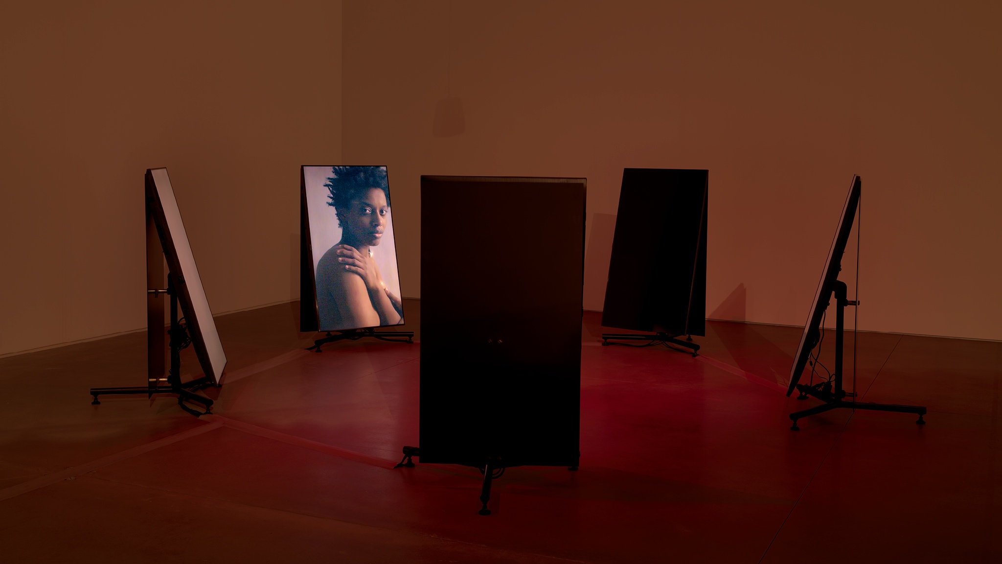 A circle of five video screens standing upright and facing inward toward each other in a gallery space. The space is lit with a diffuse red light and on one of the screens the artist Le'Andra LeSeur looks out of the video frame over her shoulder, as if at the camera.