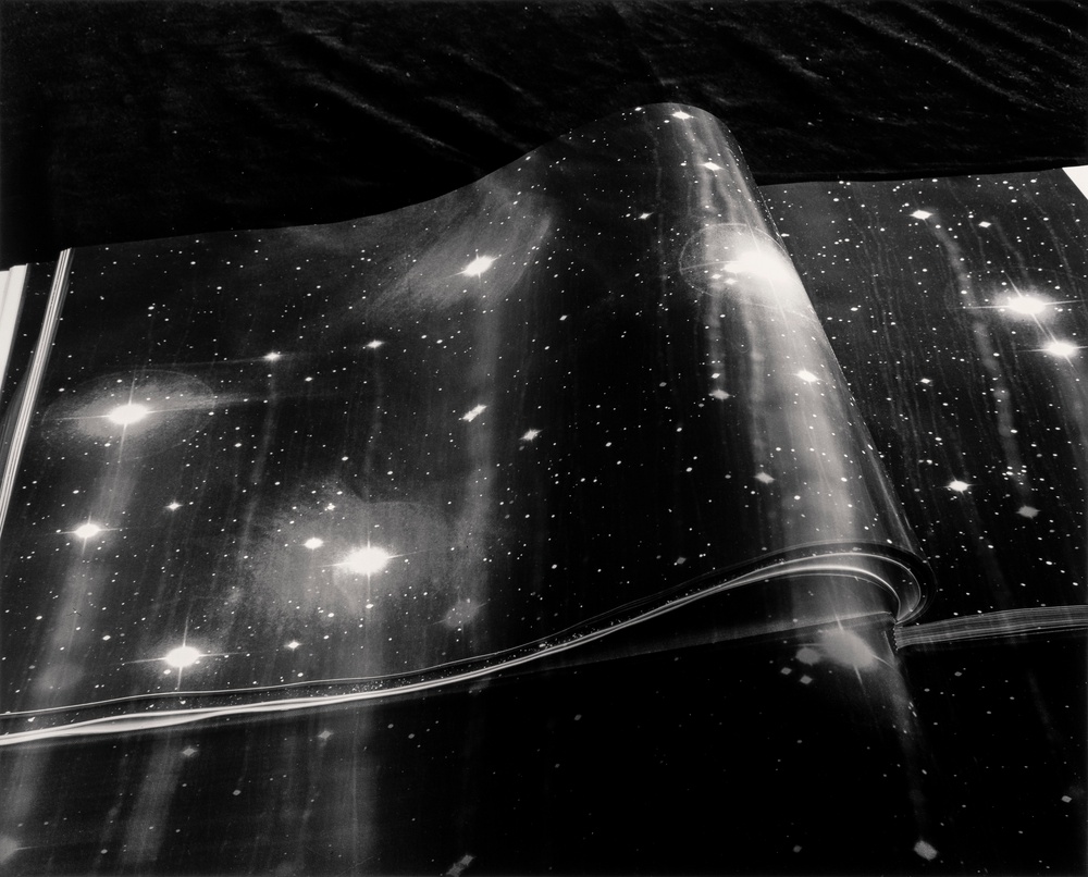 A black-and-white photograph depicting      book’s zoomed-in images of a galaxy with light beams and orbs emitting from larger stars and dotted speckles from smaller ones.