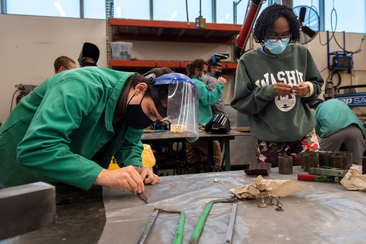 Students stand around a metal shop worktable inspecting small items in their hands. On the table sit metal saws and three small metal figurines.