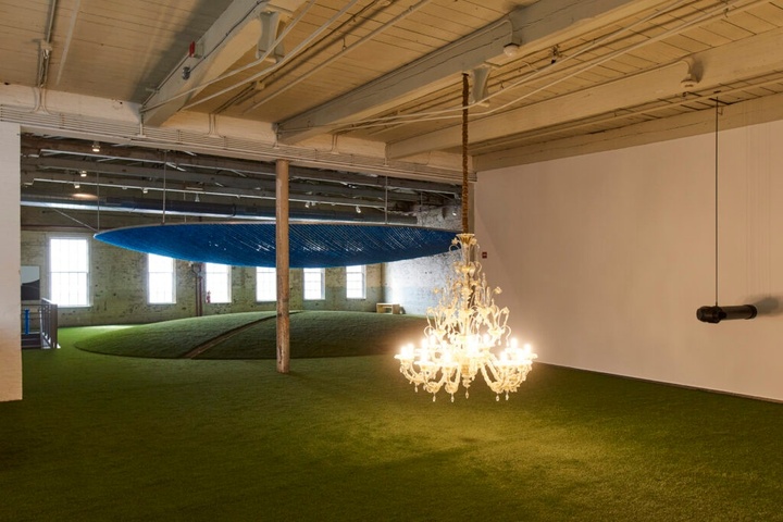 A chandelier hangs low in a gallery almost touching the ground. In the background, a green hill resembling grass with a large circular blue yarn installed above it