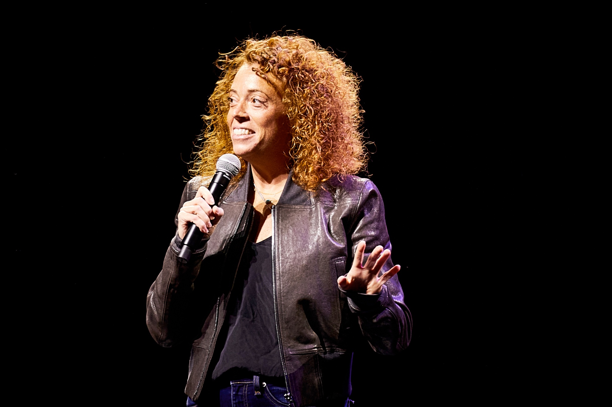 A white woman with curly red hair stands on a dark stage and holds a microphone to her mouth, looking off to the side as if at an audience 