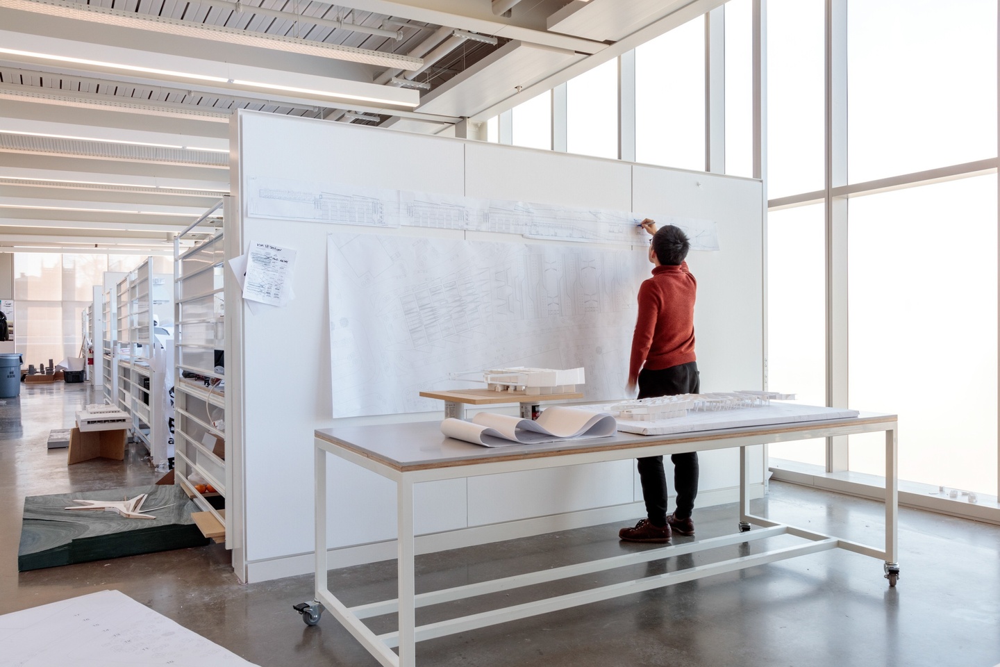 A student in a light-filled architecture studio pins up work on a white board. A table in front of the pinup board hold architectural models.