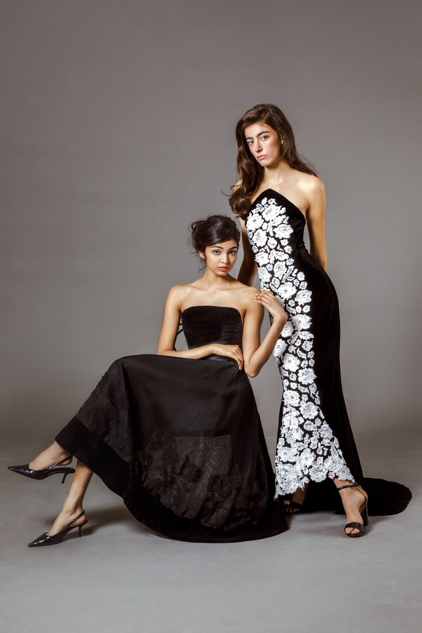 two models, one sitting in a strapless black dress and one standing in a black dress with white lace details