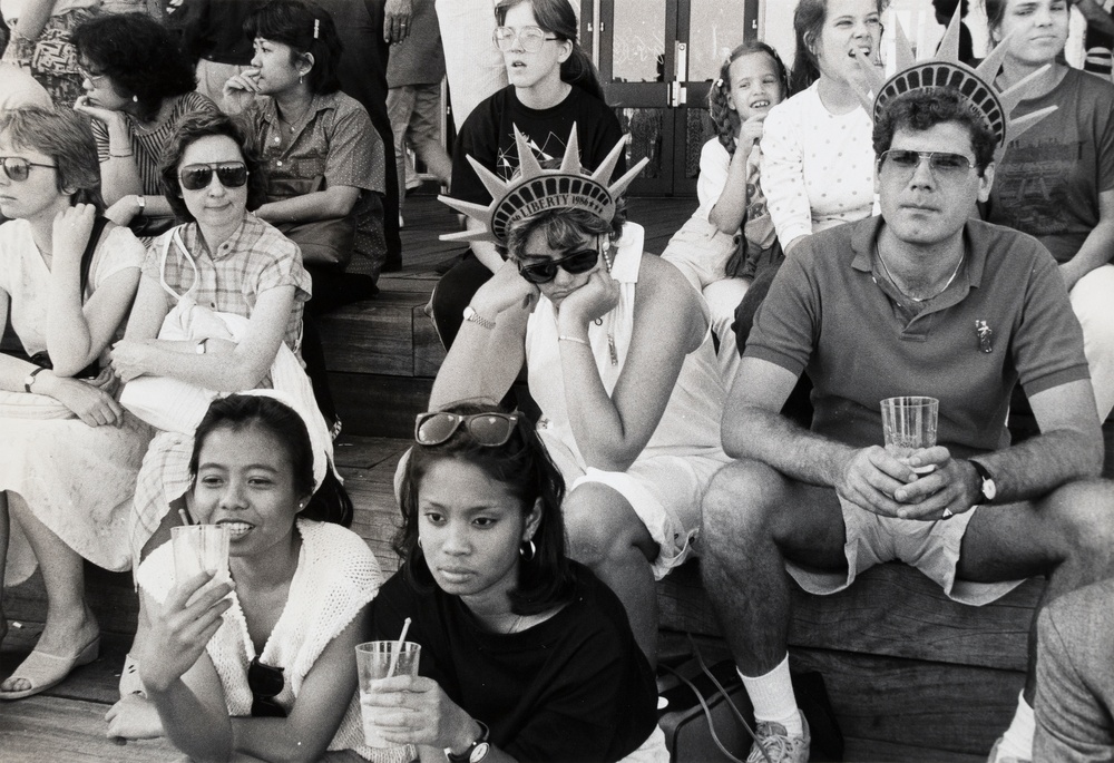 A black and white photograph of a group of people sitting on bleachers, two people seated in the center wear foam statue of liberty crowns.