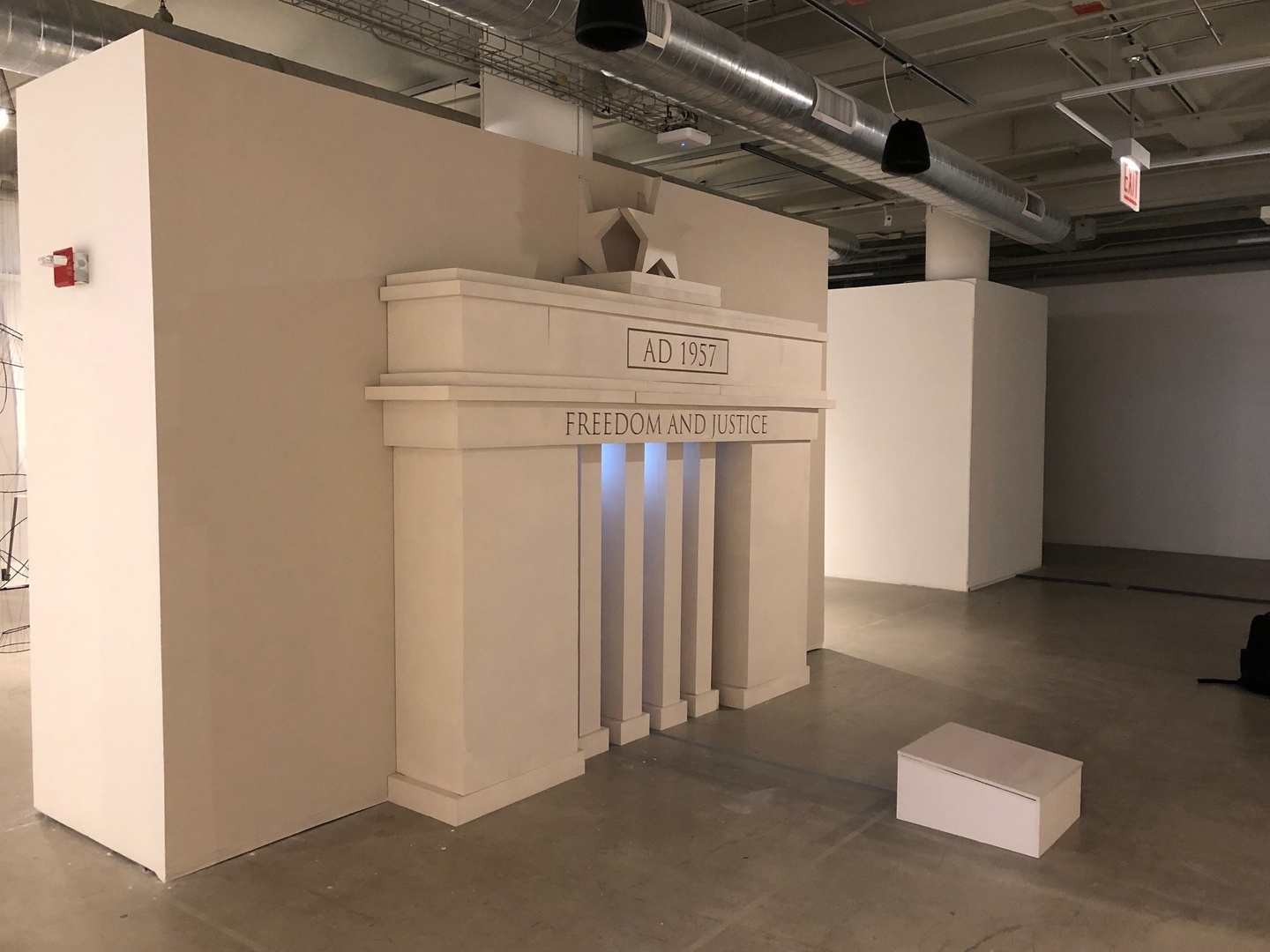 Sculptural installation set against a white wall, featuring a white building entrance, with skinny white pillars in the front opening. The inscription AD 1957 is at the top, and under it, Freedom and Justice.
